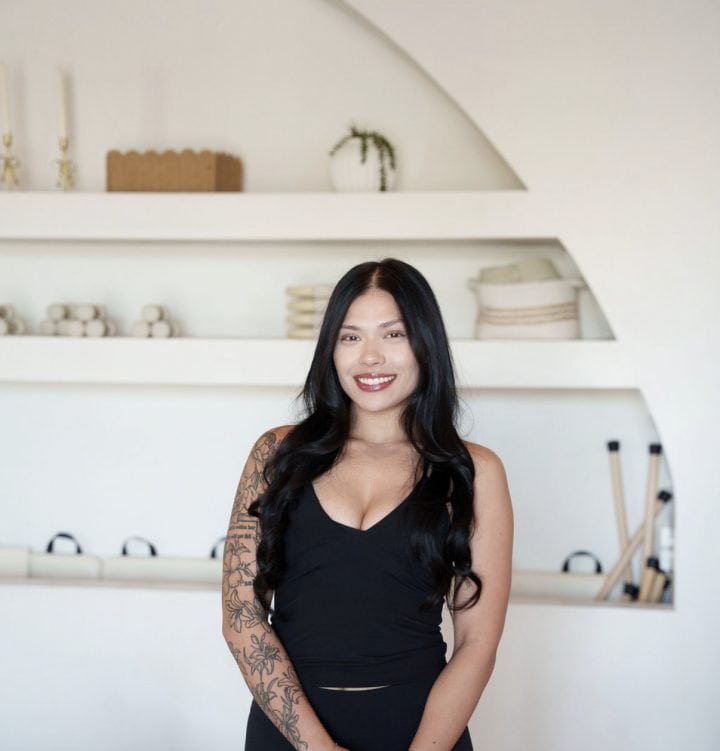 Woman with long black hair and a floral arm tattoo stands smiling in front of white shelves with ceramics and decor items.