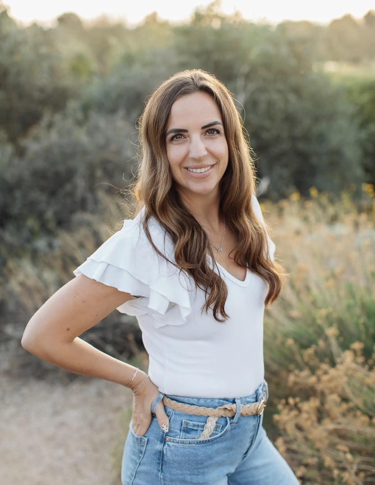 Woman with long brown hair wearing a white top and blue jeans stands outdoors, smiling at the camera with greenery in the background.