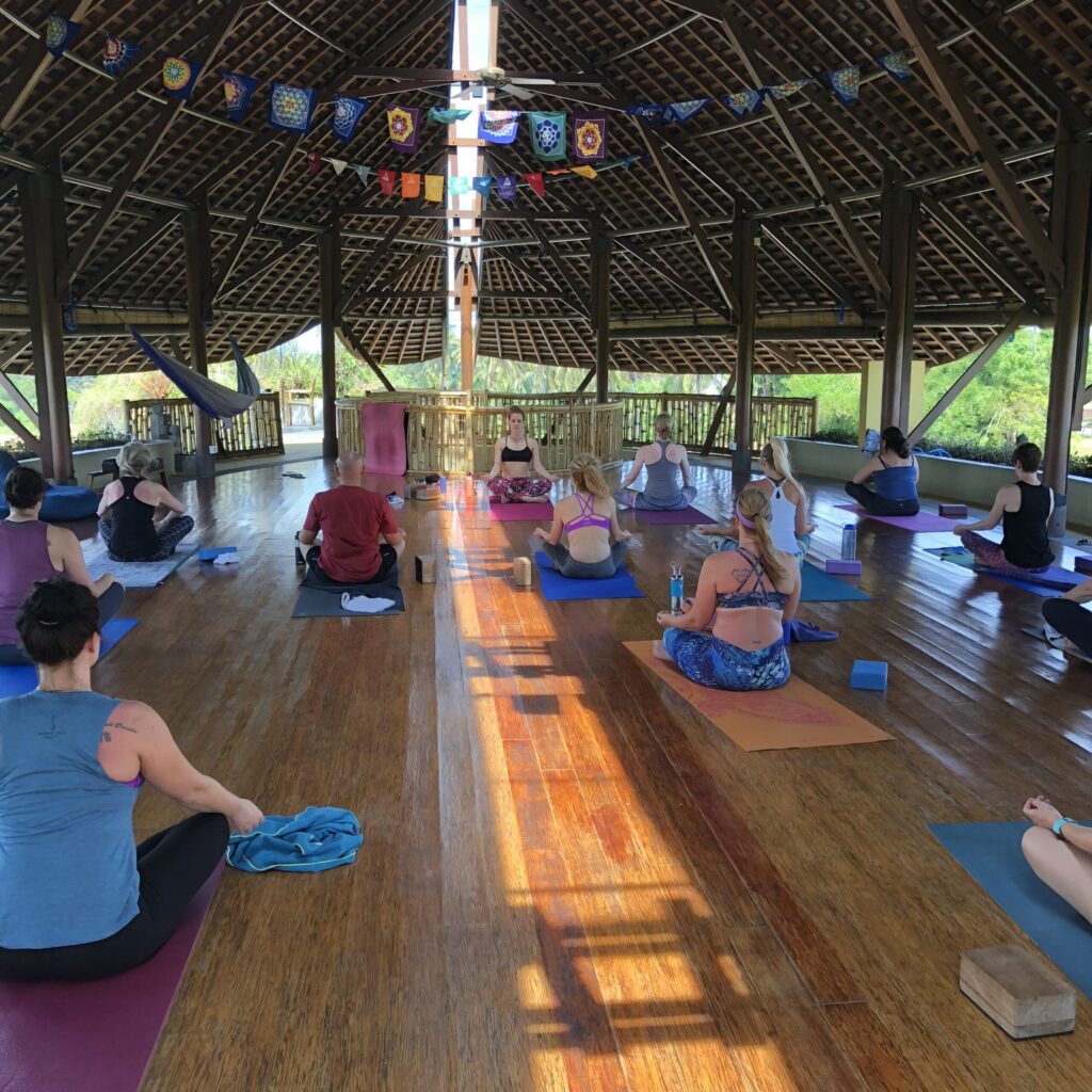People sitting on yoga mats in a large, open-air wooden pavilion with a high ceiling and decorative banners.