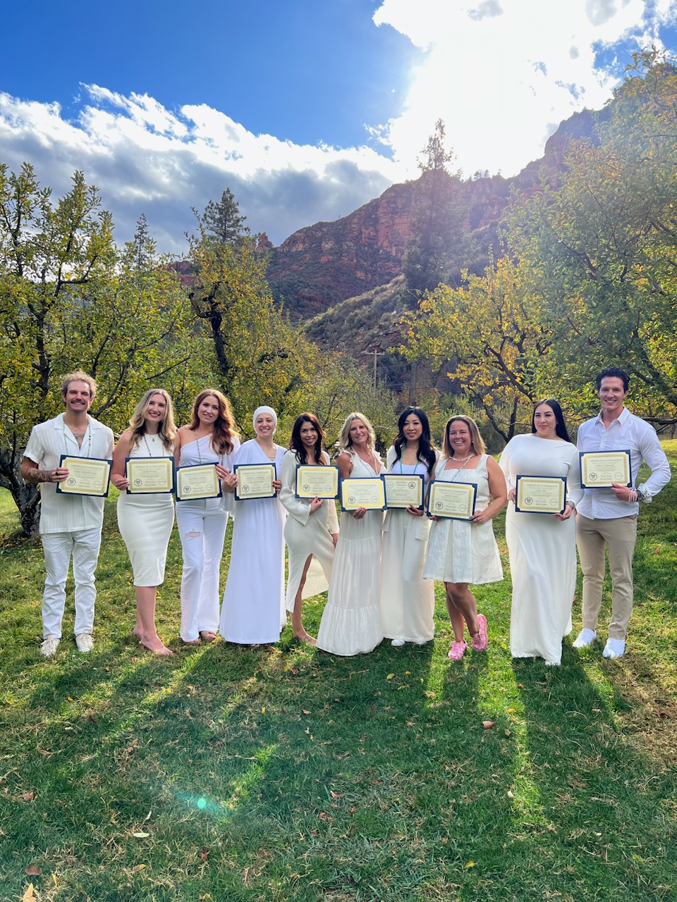 A group of ten people in white outfits stands outdoors in a grassy area with trees, each holding a framed certificate. Mountains and a cloudy sky are in the background.