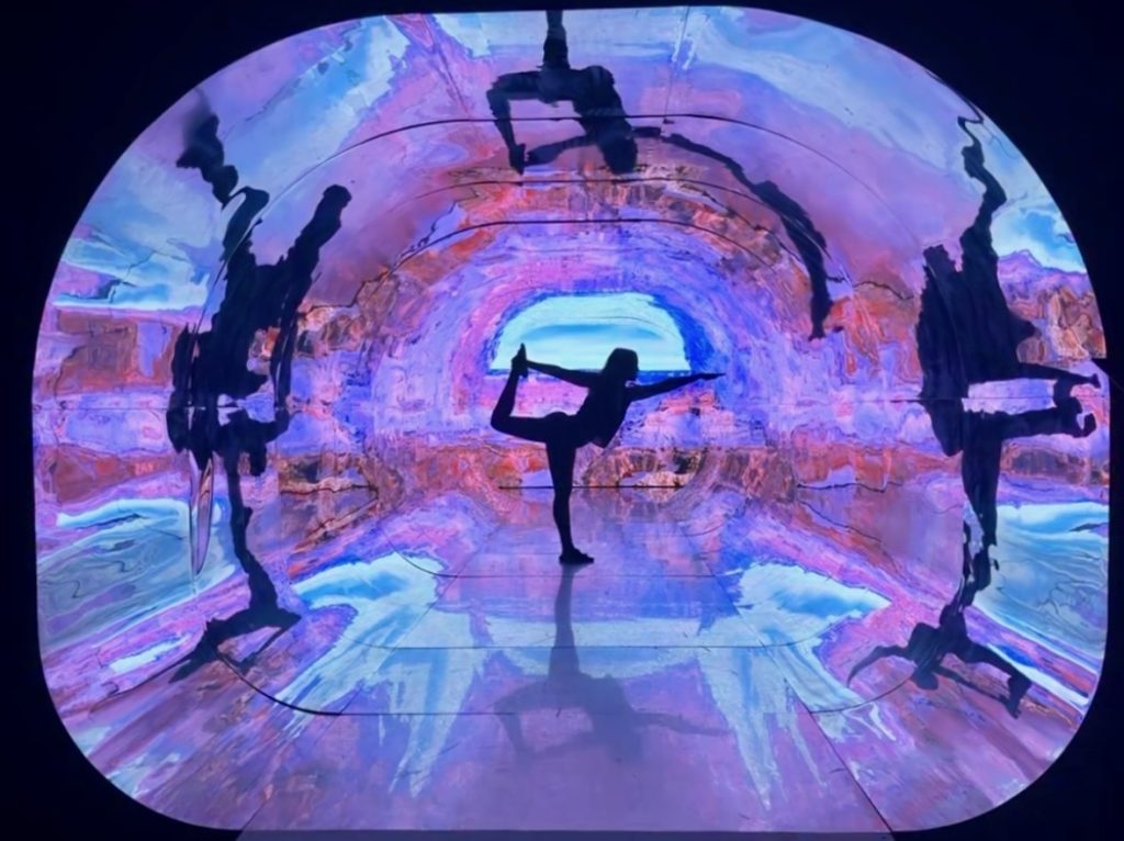 A person balances on one leg in a yoga pose inside a tunnel with colorful, abstract projected lights and reflections.