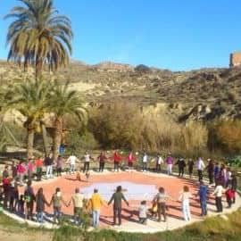 People holding hands in a circle at an eco village event with palm trees and hills in the background.
