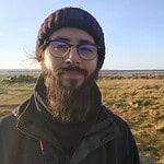 Person with beard and glasses in open landscape at sunset.