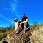 Person sitting on a rock with a dog in a natural landscape under a blue sky.