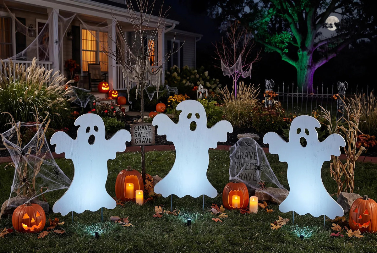 Lighted ghost cutouts with pumpkins and tombstones in a spooky yard scene.