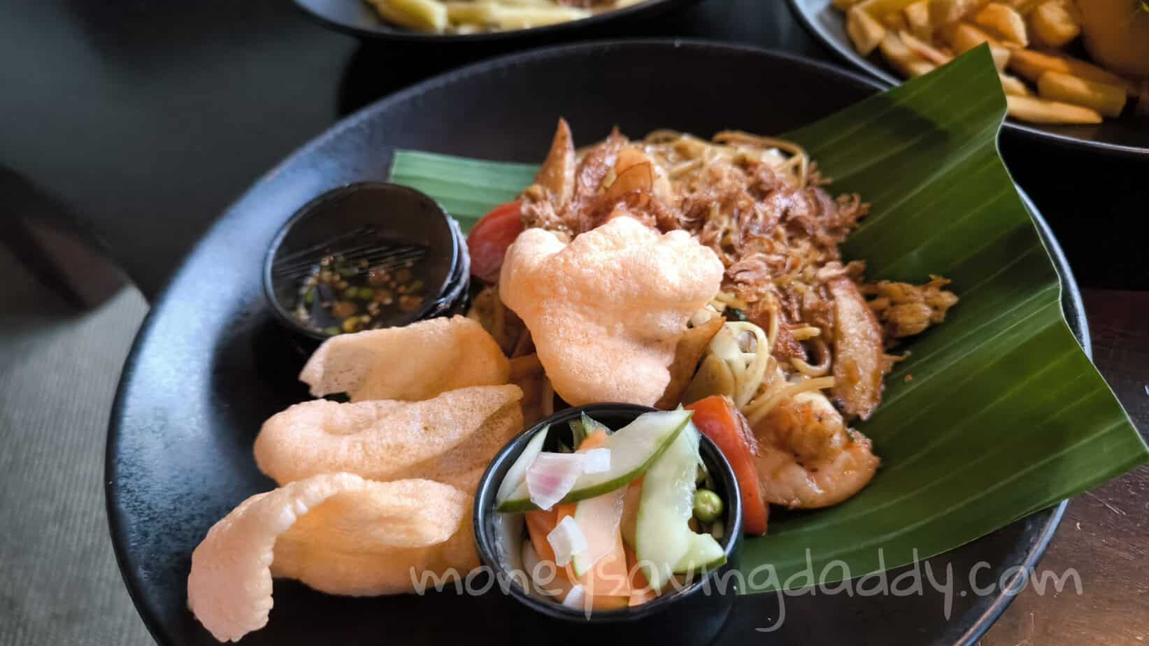 Crispy fried pork rinds with noodles and vegetables on a black plate, served with dipping sauce.