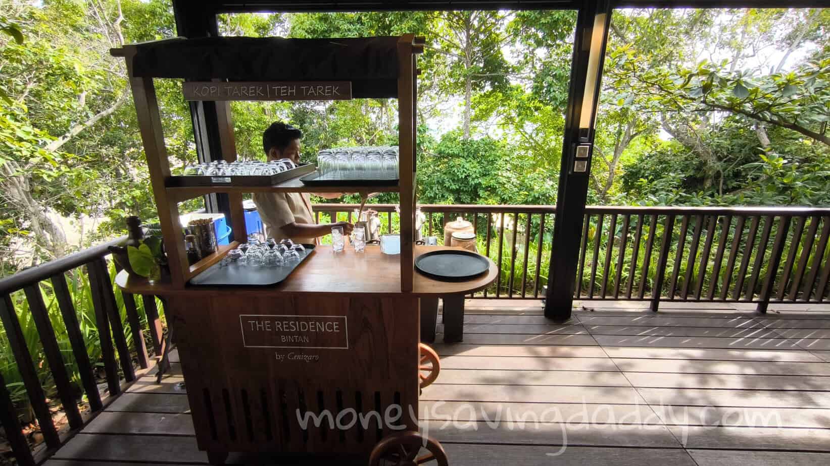 A cozy outdoor wooden bar with a person preparing drinks amidst lush green trees.
