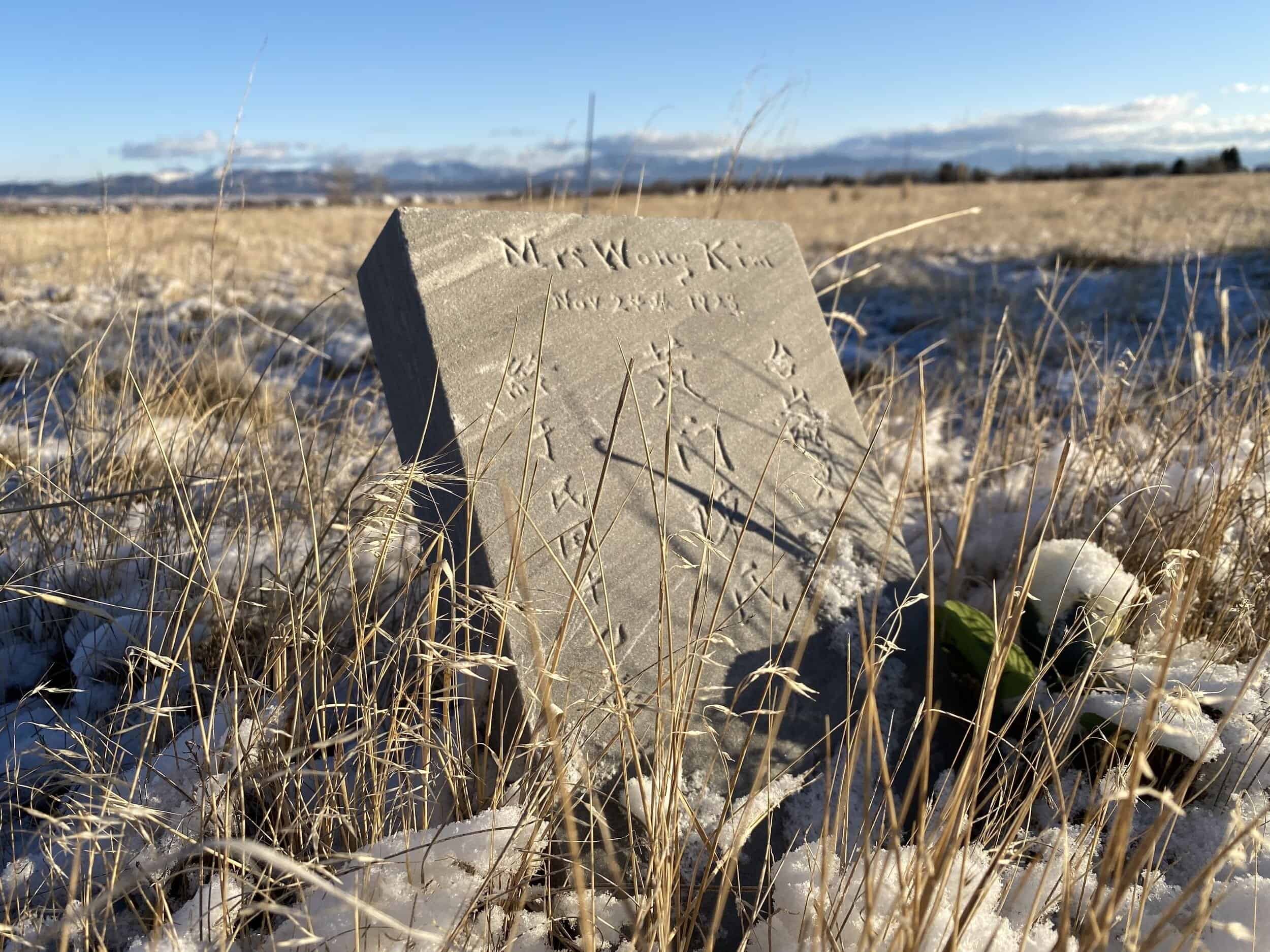 A weathered grave marker bearing Chinese inscriptions stands amidst snow-covered fields, marking the resting place of Mrs. Wong Kim—a testament to the enduring legacy of Montana’s Chinese pioneers