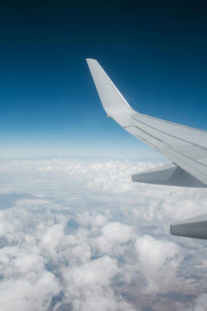 View from an airplane window showing the wing above a sea of clouds against a clear blue sky.