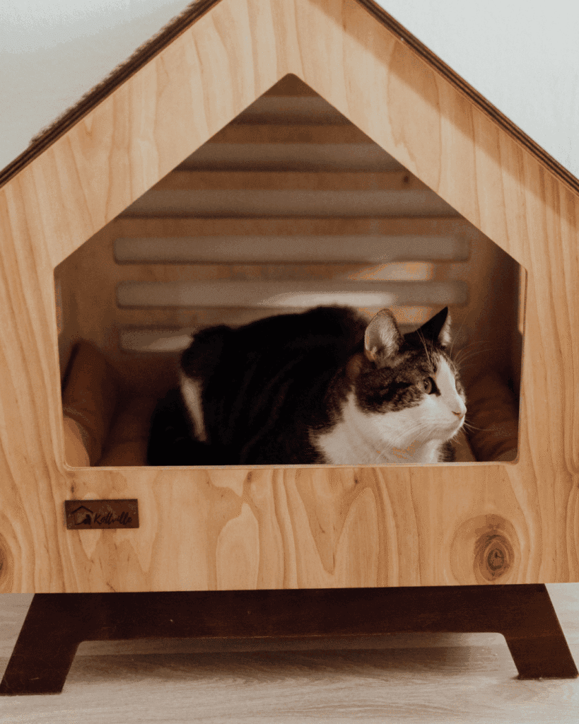 Black-and-white tabby cat curled up inside a wooden, house-shaped cat bed with a triangular roof, resting on cushions.