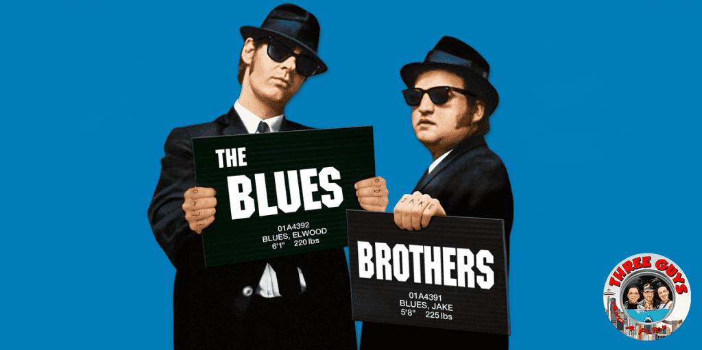 Two men in black suits and sunglasses holding movie-themed signs for "The Blues" and "Brothers".
