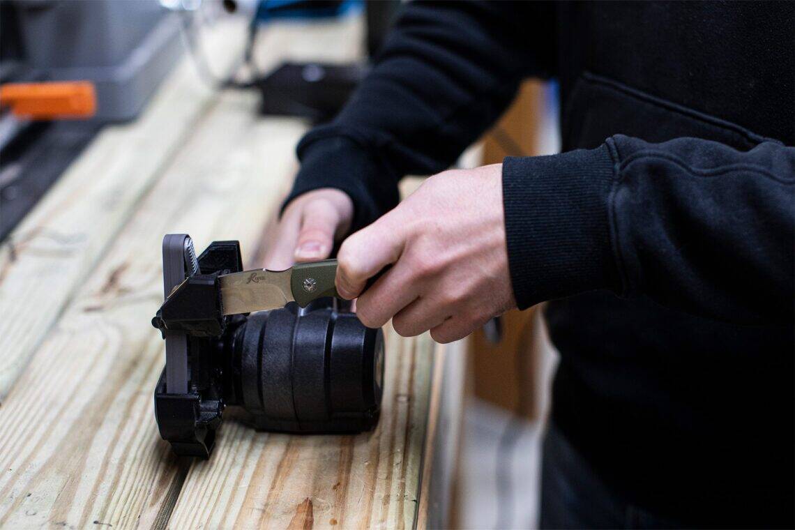 Professional electric belt sharpener being used on a Roper folding knife with an olive green handle. Essential blade maintenance tool from ABKT.