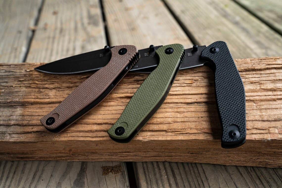 Three ABKT folding knives with D2 steel blades in tan, olive drab green, and black textured handles, resting on a wooden beam.