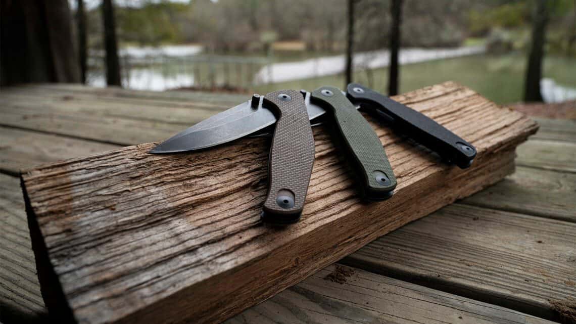 Collection of ABKT folding knives with G10 handles in brown, olive drab, and black. Features a stonewashed drop point blade resting on a wood beam.