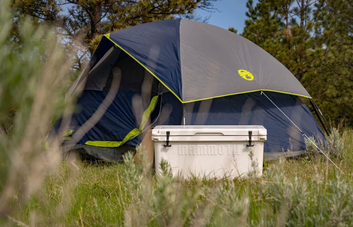 White Mammoth heavy-duty cooler sitting in front of a blue and grey camping tent in a grassy outdoor field.