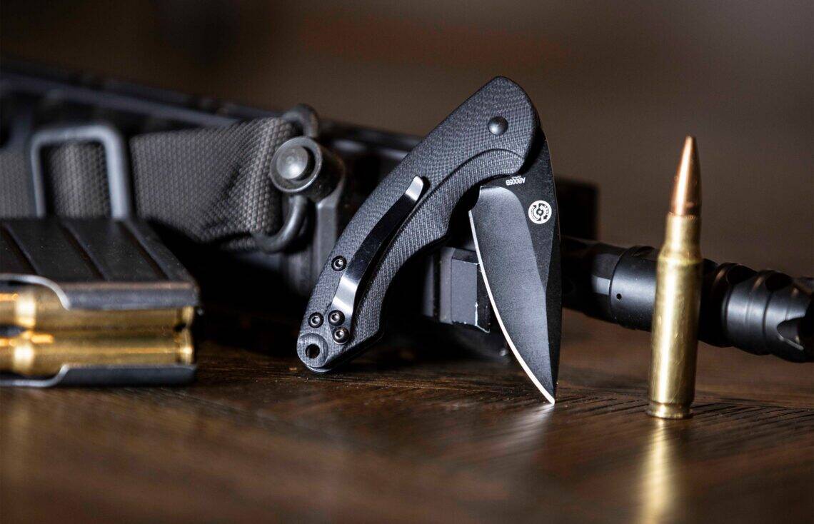 All-black ABKT tactical folding knife with a textured G10 handle and black-coated blade, displayed with a rifle and magazine.