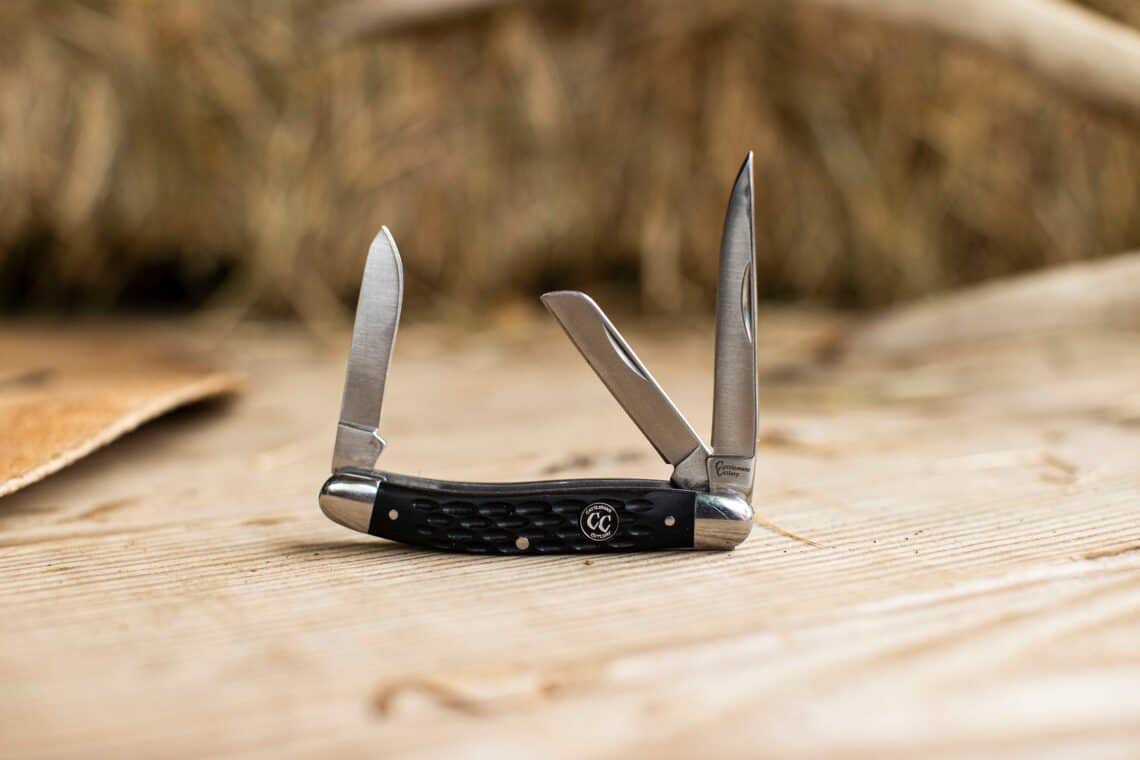 A traditional Cattleman's Cutlery folding knife with three blades and a black jigged bone handle, open on a weathered wood surface.
