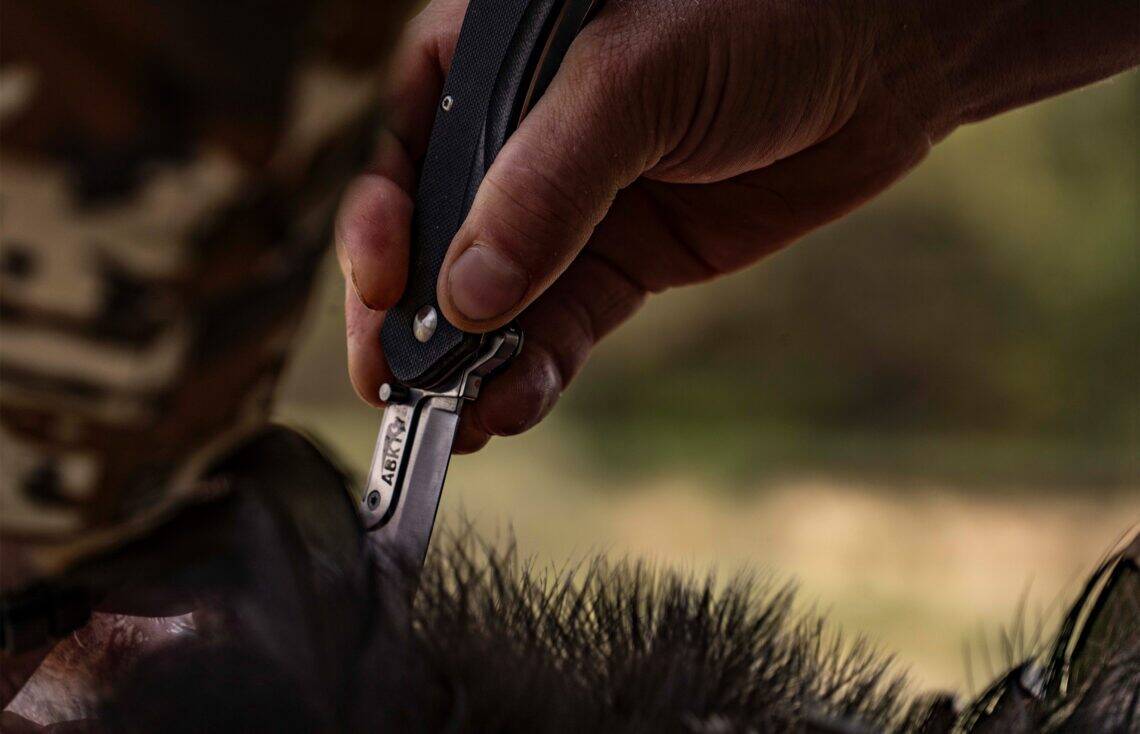 ABKT tactical folding knife with a black G10 handle and stainless steel blade being used outdoors. Professional hunting and skinning tool.