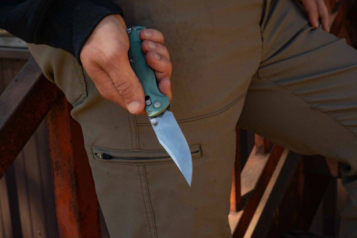 Close-up of a hand holding an ABKT folding knife with an olive green handle, featuring a crossbar lock and drop point blade.