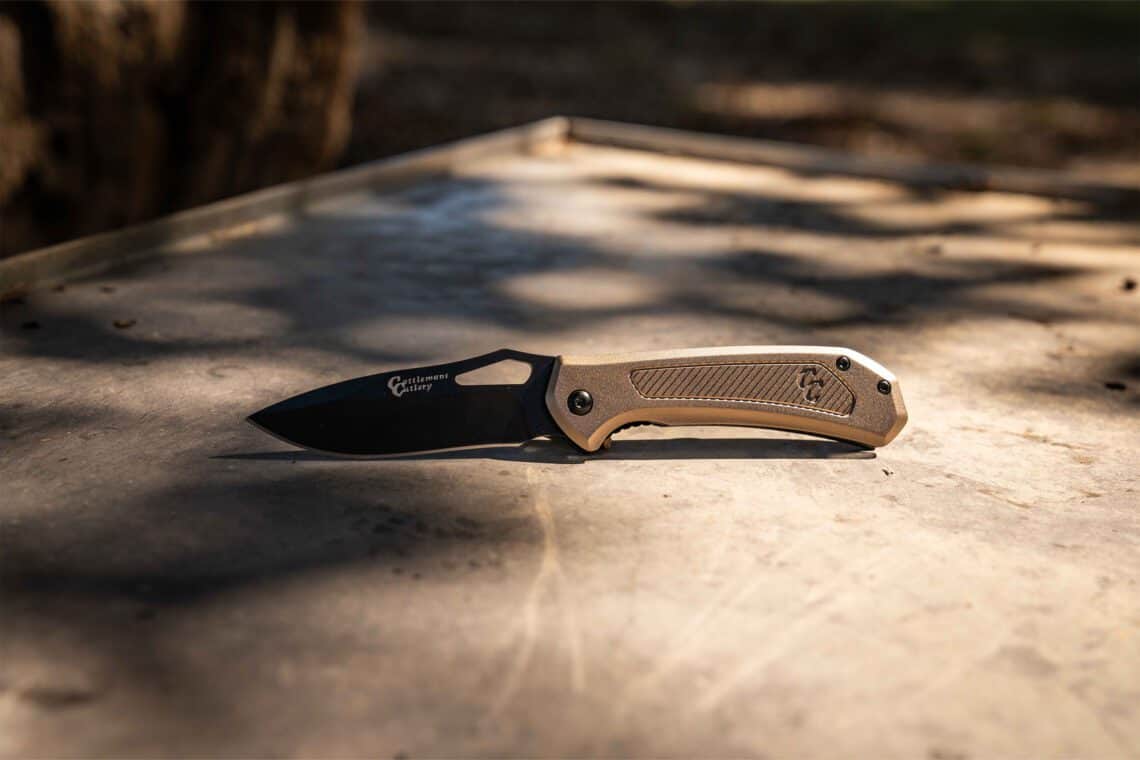 Cattleman's Cutlery folding knife with a black coated blade and a champagne gold aluminum handle on a rustic wooden surface.