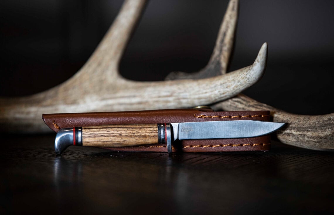 ABKT fixed blade hunting knife with a stacked wood handle and leather sheath, displayed in front of a deer antler.