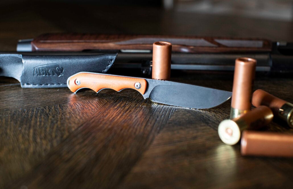 ABKT EDC folding knife with a black textured handle and cleaver-style blade, lying on a wooden table next to a rifle magazine and a brass casing.