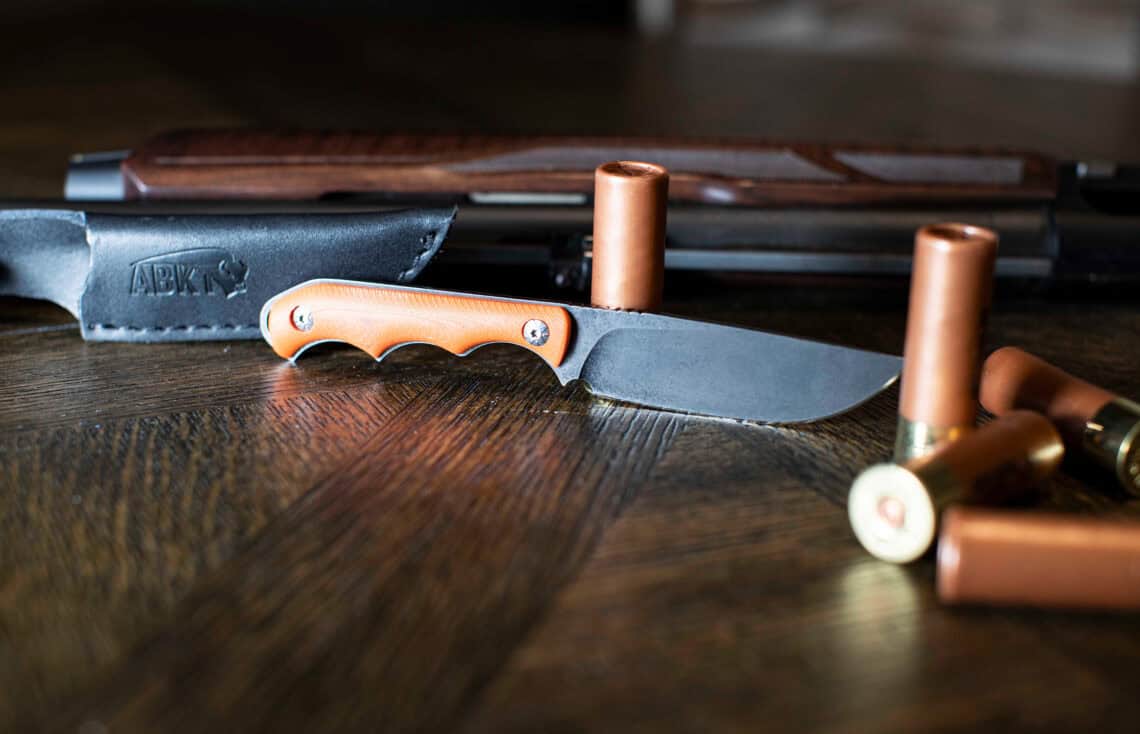 ABKT EDC folding knife with a black textured handle and cleaver-style blade, lying on a wooden table next to a rifle magazine and a brass casing.