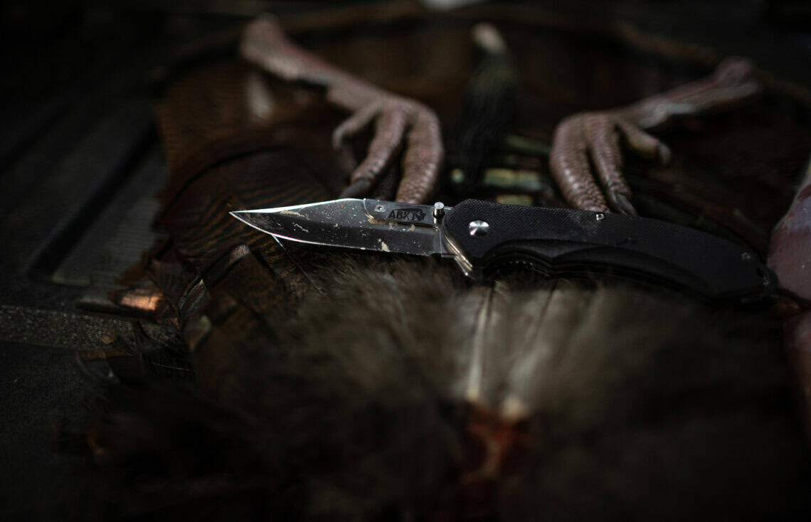 Close-up of an ABKT tactical pocket knife with a black handle and sharp blade used in a hunting scenario. Professional-grade outdoor equipment.