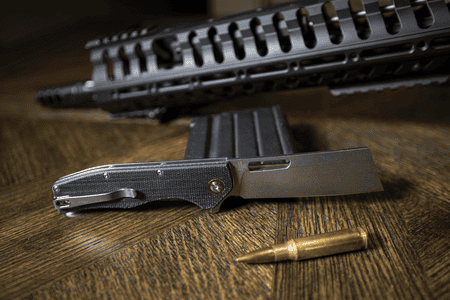 ABKT EDC folding knife with black textured handle and cleaver-style blade, resting on wood with a rifle and round.