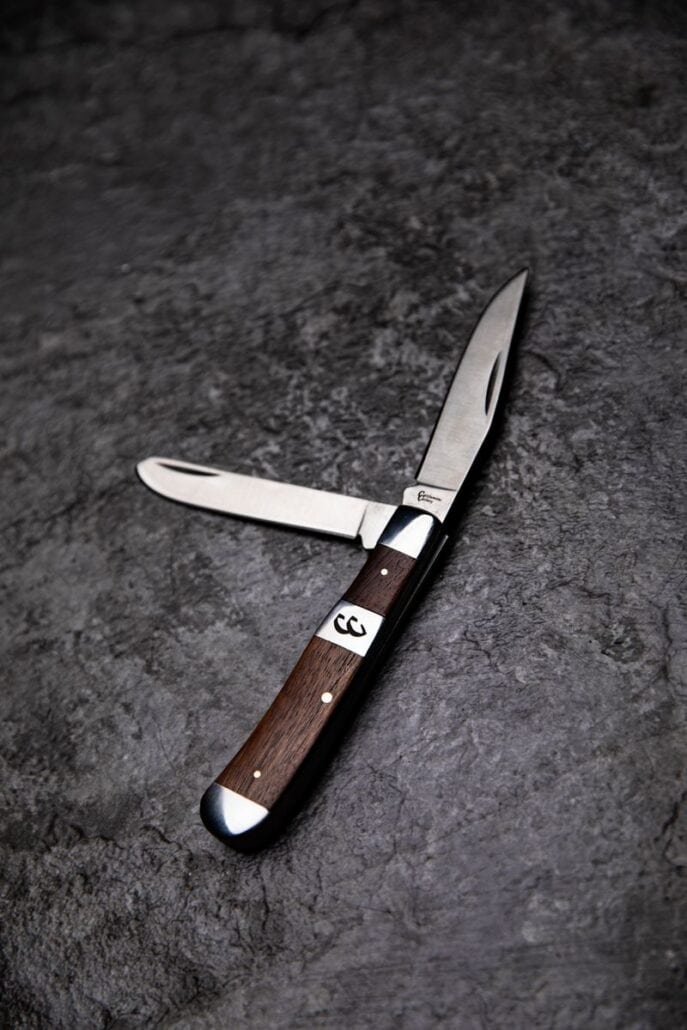 A Cattleman's Cutlery two-blade folding pocket knife with a dark wood handle and dual bolsters, resting on a textured dark surface.