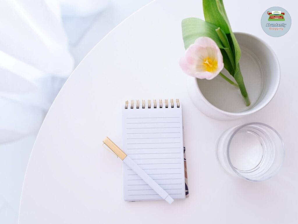 flatlay of a little notebook with pen on a white round table - glass of water and a flower in a vase - benefits of goal setting