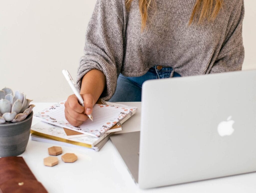 woman behind her laptop, writing in her notebook in front of her - how to blog consistently