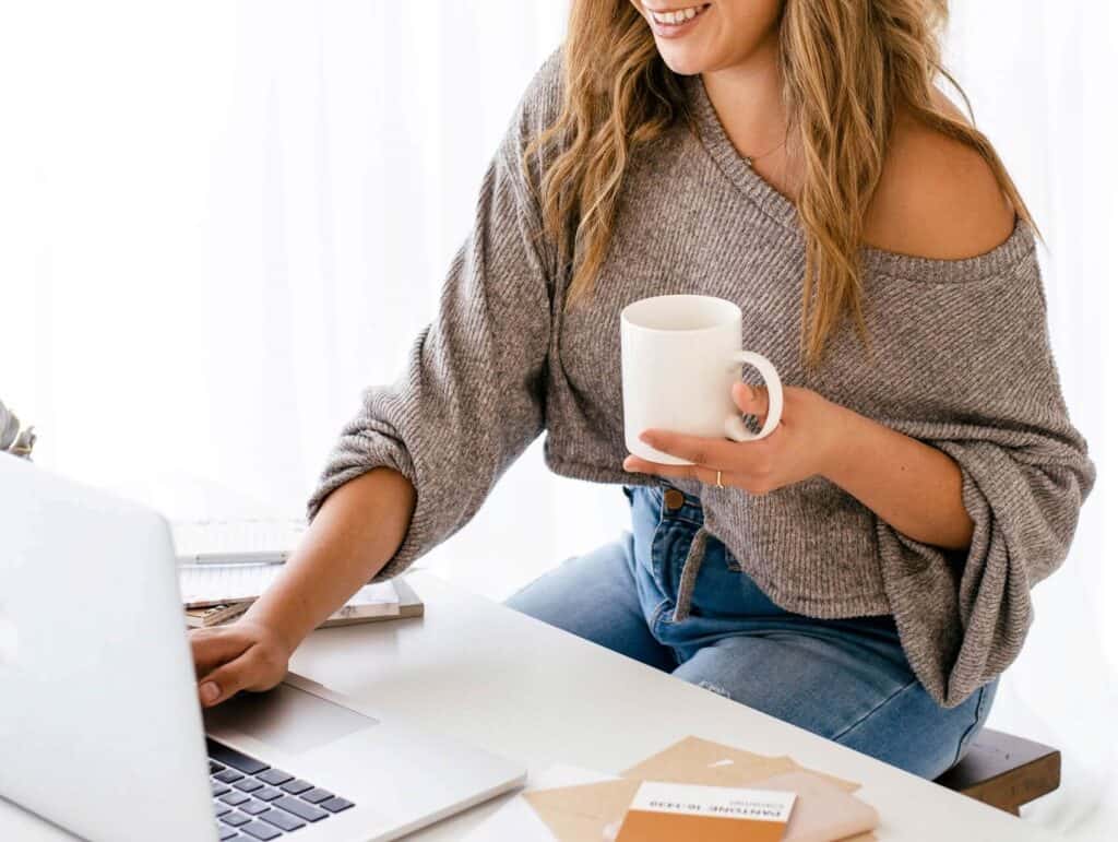 woman behind her laptop, holding a mug, notebooks in front of her - how to blog consistently