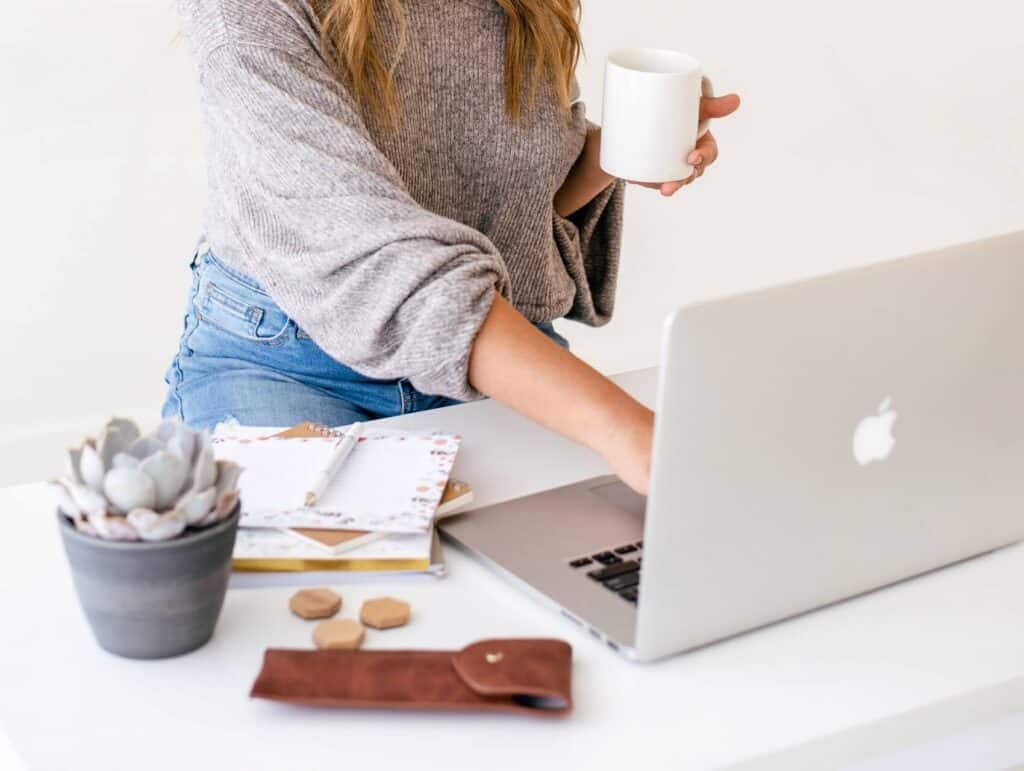 woman behind her laptop, holding a mug, notebooks in front of her - how to blog consistently