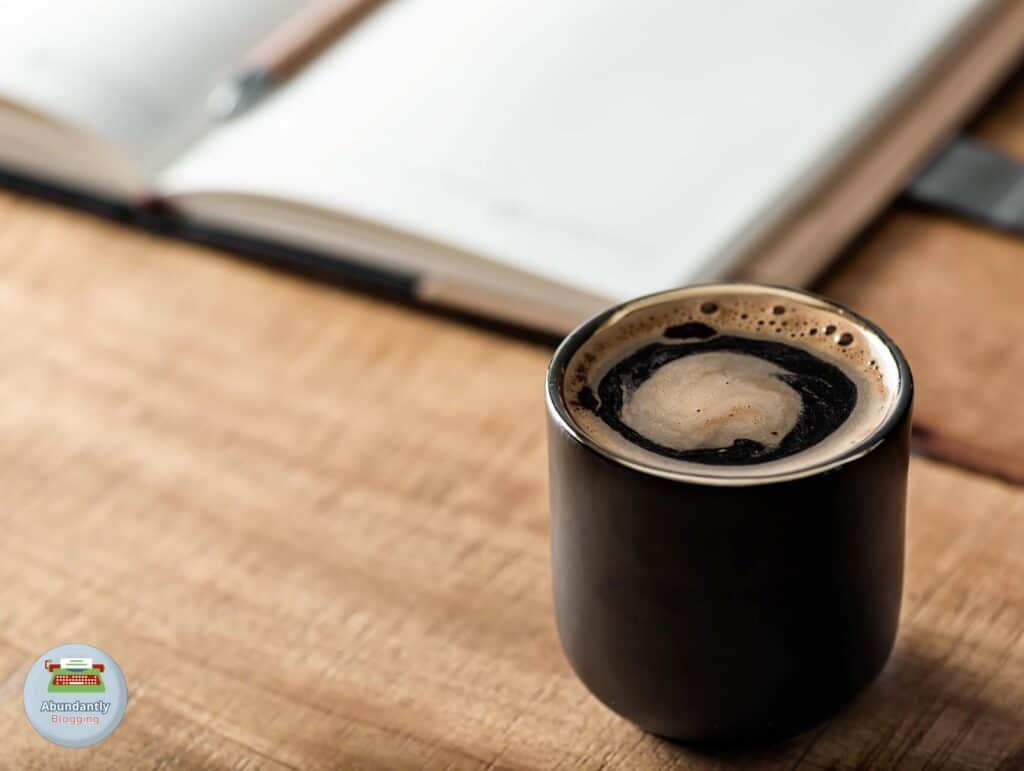 A wooden desk, with a open notebook and pen and on the foreground a black mug with coffee - Reasons for Procrastination