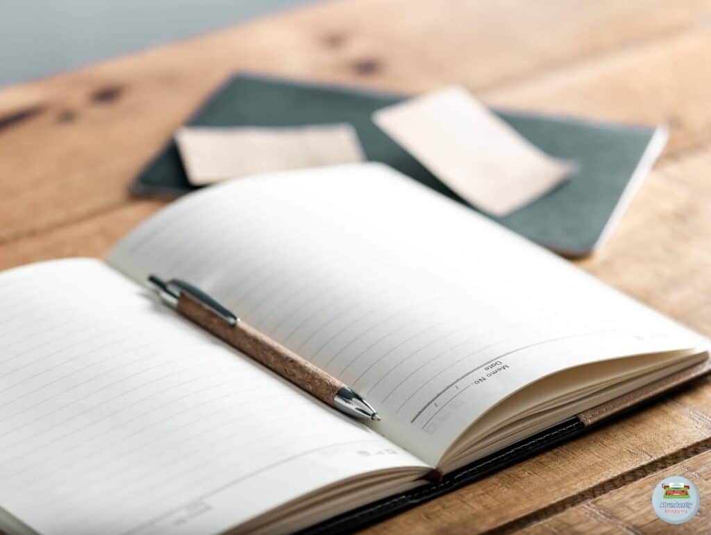On a wooden desk lays a open notebook with a pen in it. In the background there is another notebook and some post-its - Reasons for Procrastination