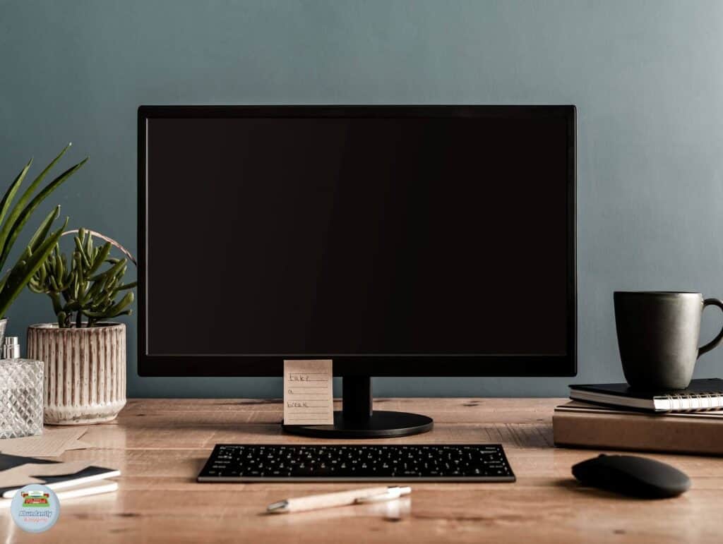A wooden desk with a computer monitor (it's off), a keyboard and mouse, several notebooks, some plants, and a pen - Reasons for Procrastination