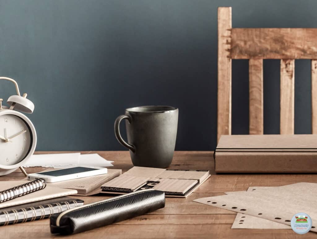 A wooden chair and desk with items like notebooks, a pen etui, a mug, and an analog alarm clock scattered around. - Reasons for Procrastination
