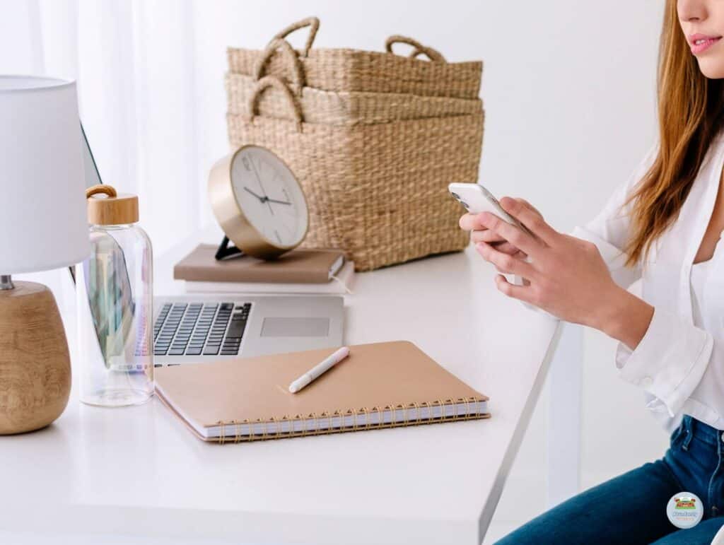 A woman working at a white desk checking her phone having her notebook and her open laptop in front of her - 80/20 Rule in Blogging