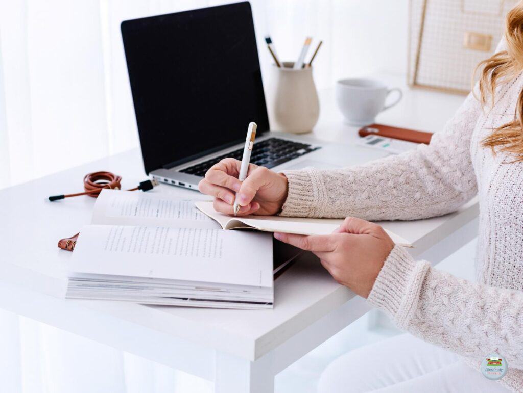 A woman working at a white desk writing in her notebook with her laptop open - 80/20 Rule in Blogging