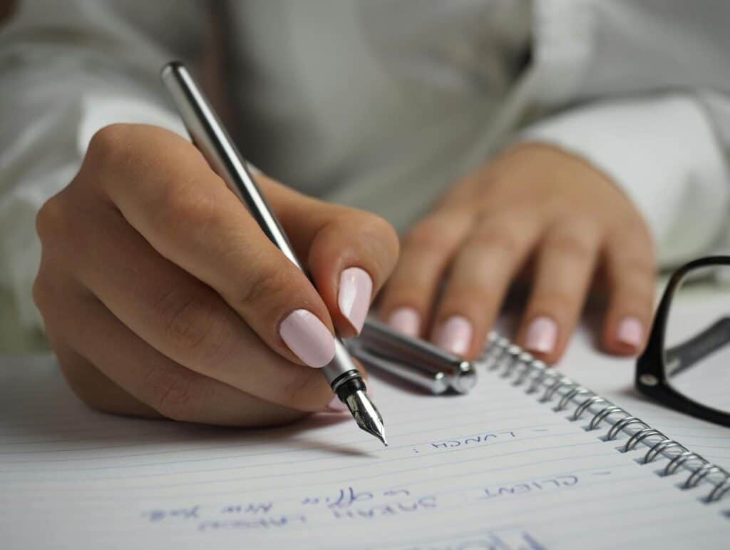 a close up of a woman writing in a notebook with a dip pen. She has pink nails and her glasses nearby - evening routine ideas