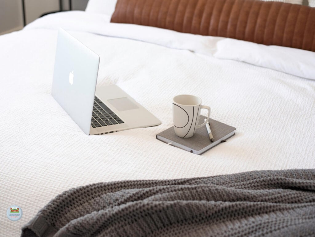 a open laptop, a notebook, pen, and mug and a knitted throw blanket on a bed with white covers - How to repurpose