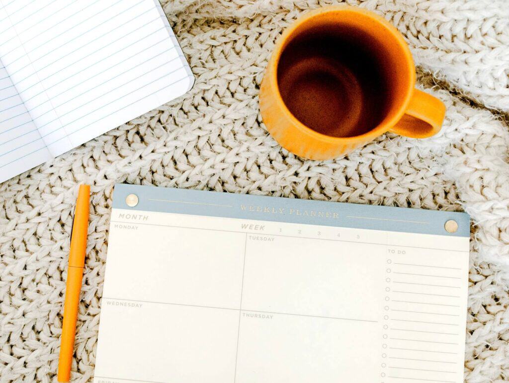 flatlay of a calendar, notebook, yellow pen and cup on a bed with a crocheted bedspread - morning routine for bloggers