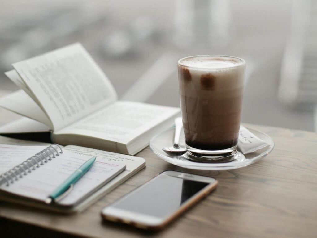 a desk with a book, some notebooks, a pen, a phone, and a glass of hot chocolate - morning routine for bloggers