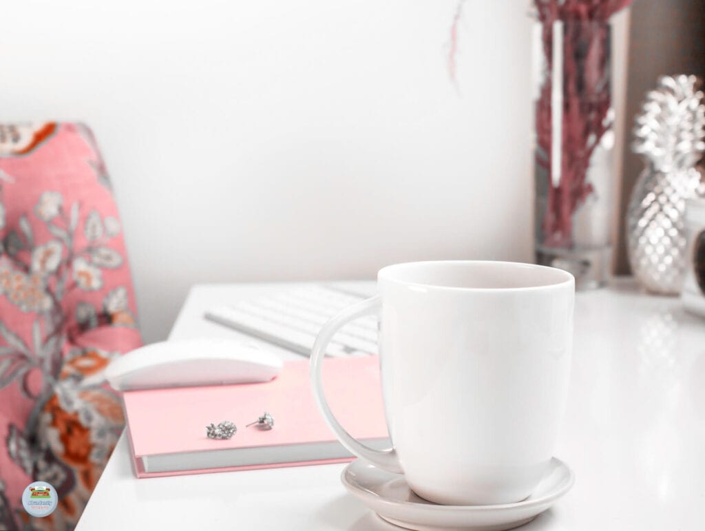 A white desk with a modern keyboard and mouse. There's a notebook, some earrings, and a cup on a saucer on the desk. By the desk is a chair in flowery pink - Setting Realistic Deadlines