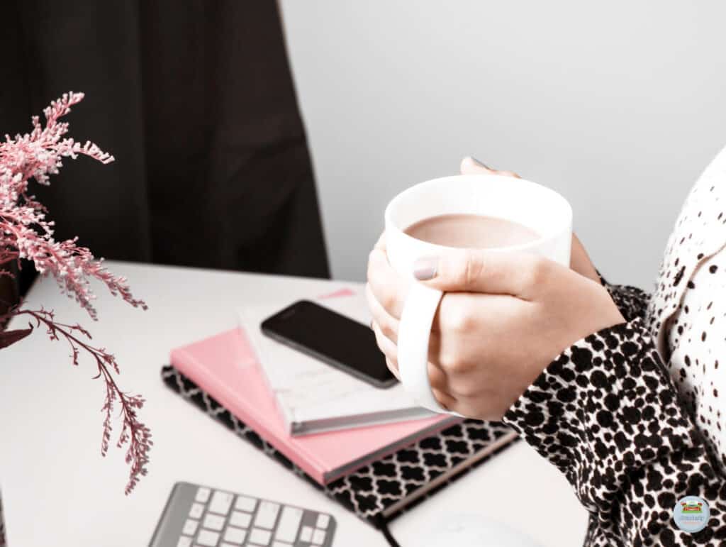 A woman holding a cup of coffee with 2 hands standing in front of a desk with a modern keyboard, some notebooks, a phone, and a pink flower - Setting Realistic Deadlines