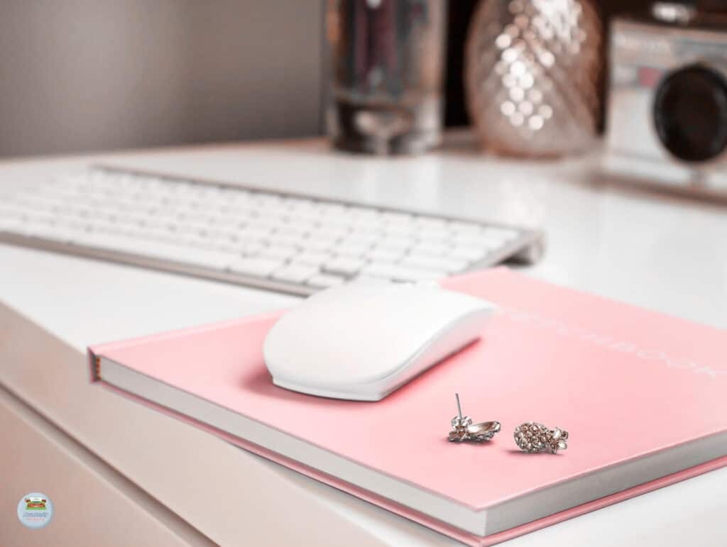 A white desk with a modern keyboard, a pink notebook with a mouse on top and some earrings - Setting Realistic Deadlines