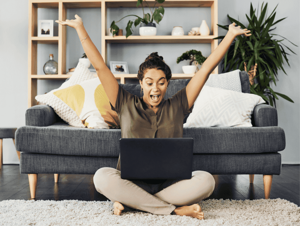 woman sitting on a rug on the ground before her grey couch with her open laptop on her lap. She is happily looking at her laptop and celebrating with her hands in the air - celebrating blogging milestones