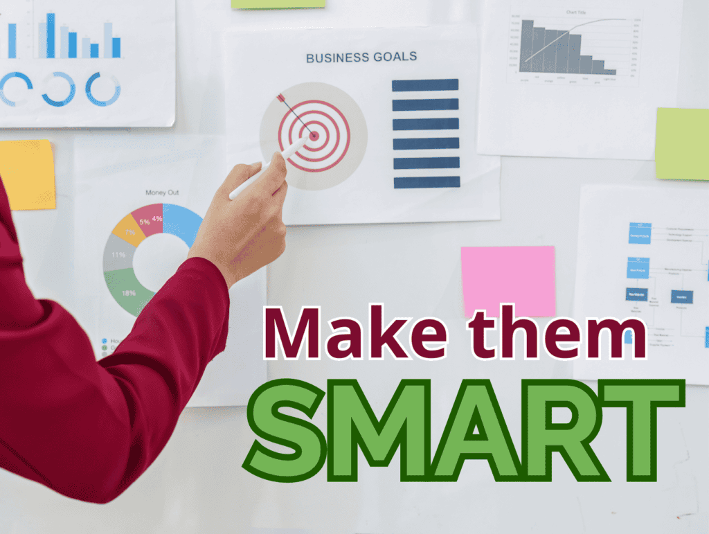 A white smart board with papers on it. On the papers are different projections and business charts. Text on the image: Make them SMART - long-term blogging goals