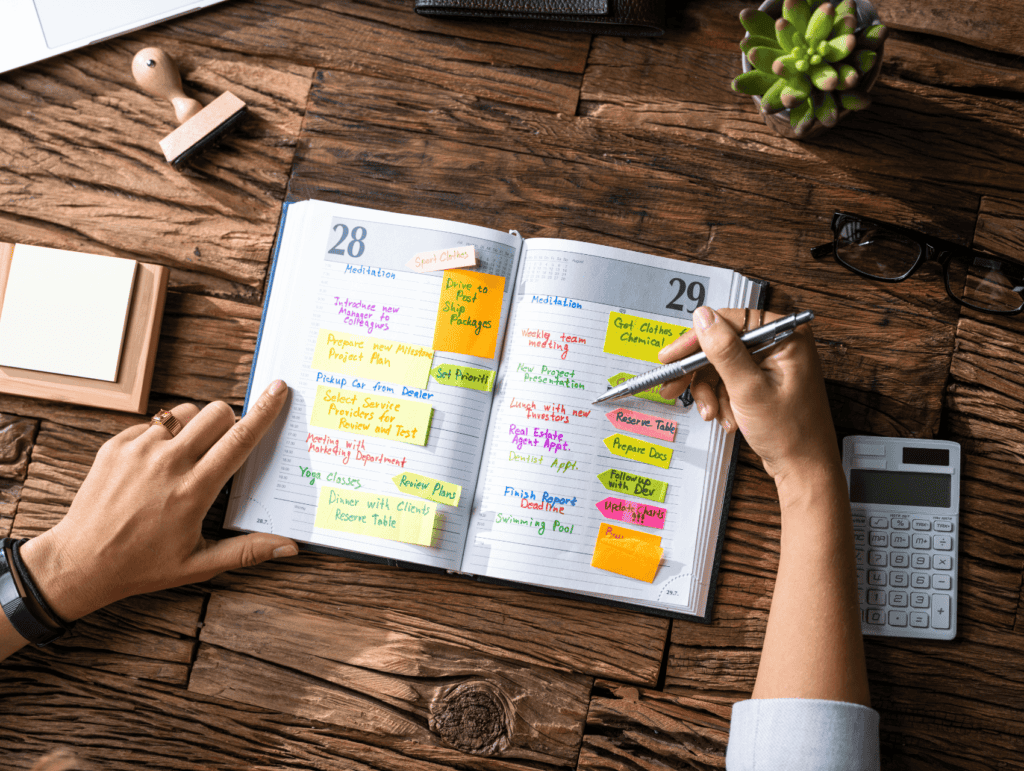 an open calendar book with lots of post-it notes in it. A woman's hands re holding it open and ready to write in it. The calendar book is laying on the table with several items surrounding it - long-term blogging goals